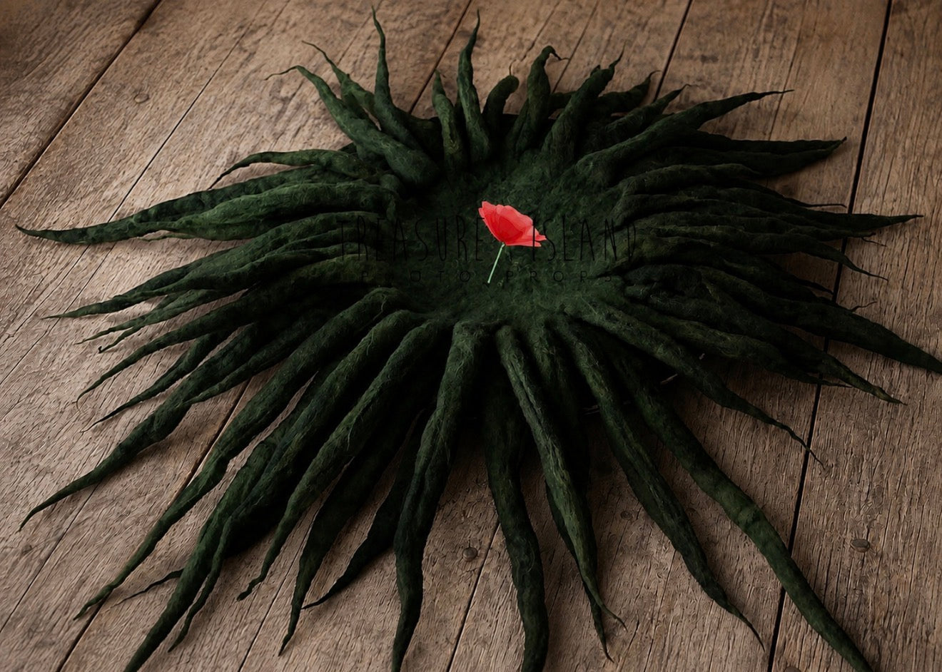 A dark green felted wool crown with long, wispy dreadlock-like strands radiating outward, displayed on a rustic wooden plank surface with a small red flower pinned in the center.