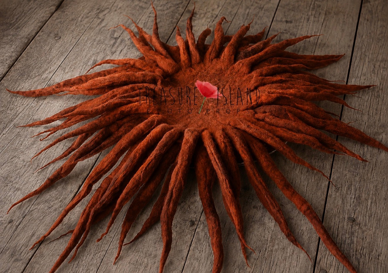 A large, circular dreadlock wreath or wall hanging made of many long rust-brown dreadlocks radiating from a central ring, with a small pink rose tucked into the middle, displayed on a weathered gray wooden plank surface.