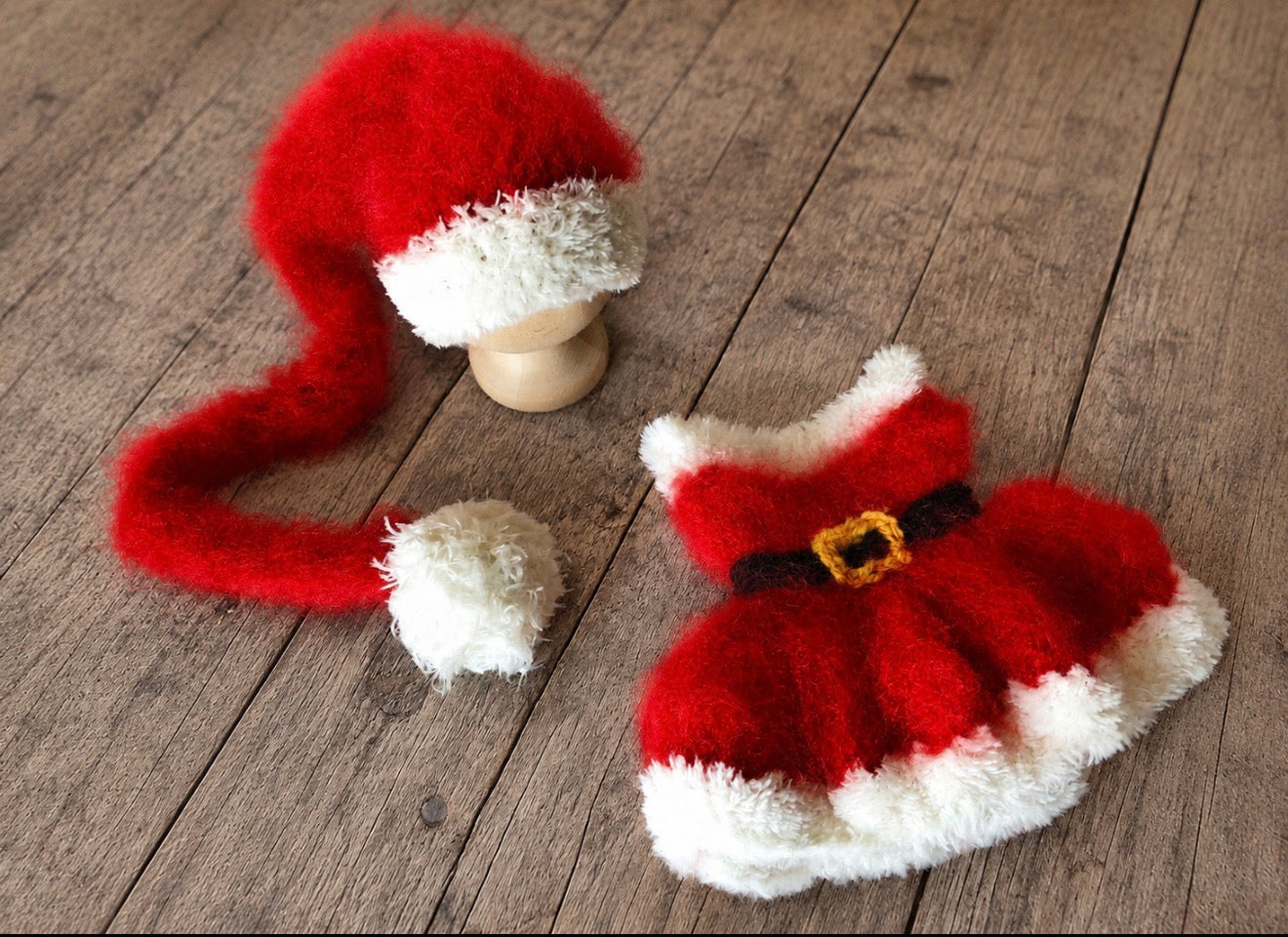 A red and white fuzzy Santa hat with a long tail and matching Santa dress with a black belt and gold buckle, displayed on a rustic wooden surface.