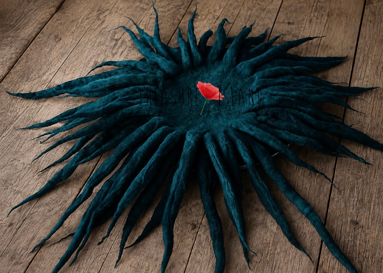 A dark teal dreadlock hat featuring long, textured synthetic dreadlocks radiating from a circular base, with a small red rose pinned in the center, displayed on a rustic wooden plank background.