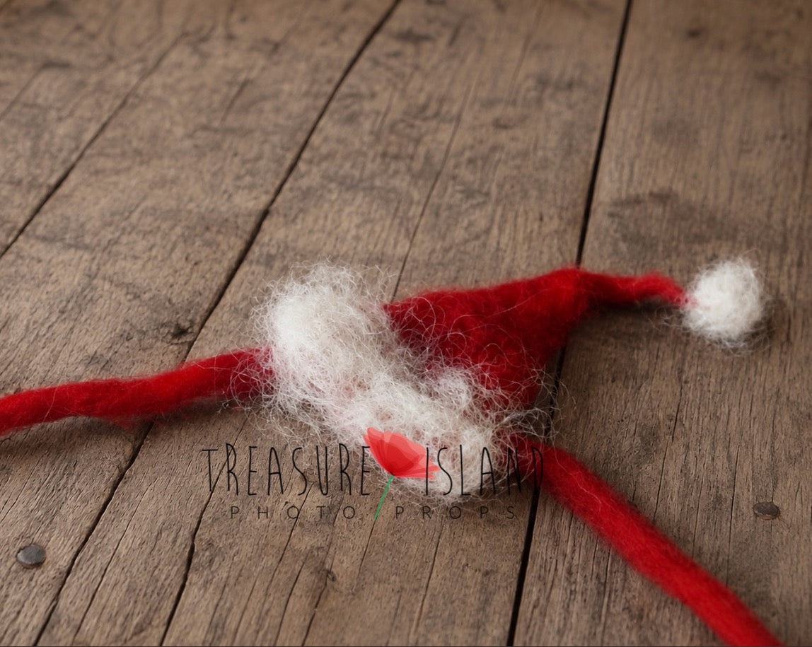 A handmade red and white felt Santa hat with messy white wool on the brim, laid flat on a rustic wooden plank surface. A watermark for 'Treasure Island Photography' is visible in the center.