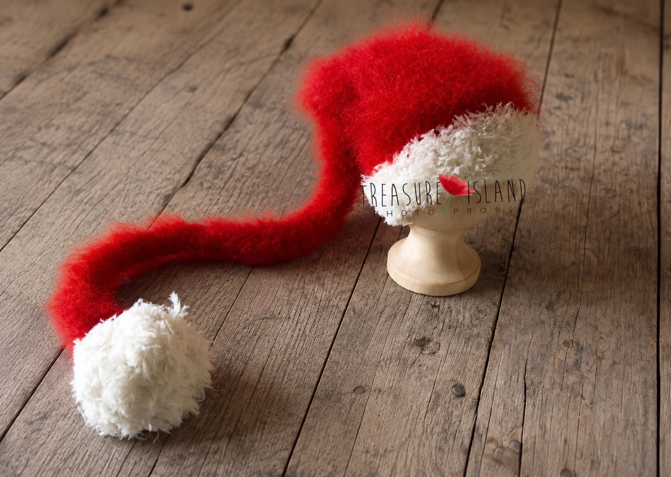 A fuzzy red and white Santa hat displayed on a small wooden stand over a rustic wooden surface. The hat has a long red tail with a white pom-pom and a white fluffy brim.