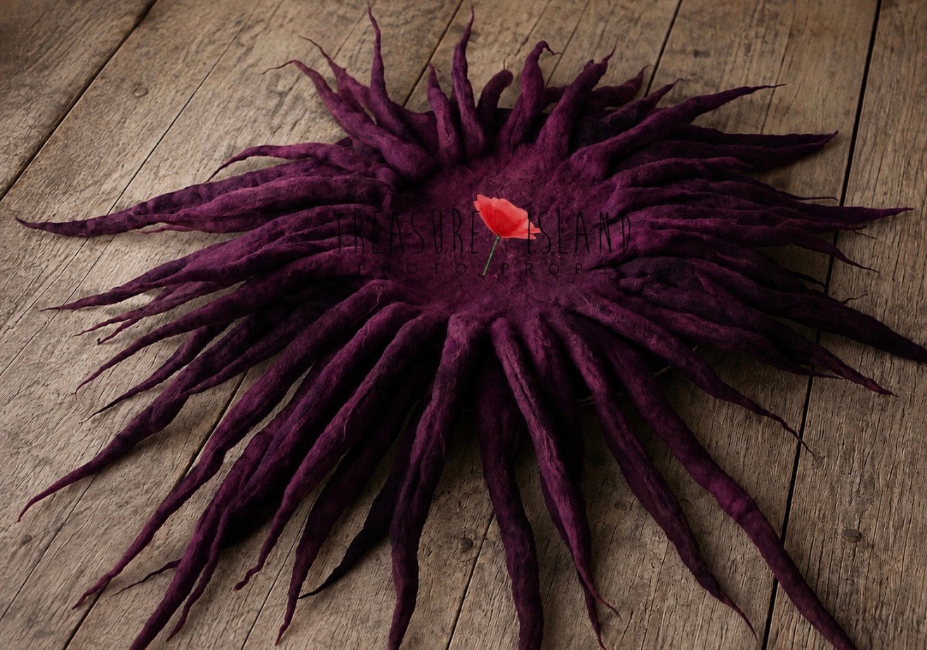 A dark purple felted wool rug with a sunburst or dreadlock-like radial pattern, photographed from above on a rustic wooden plank surface. A small red flower is placed in the center of the rug as a decorative prop.