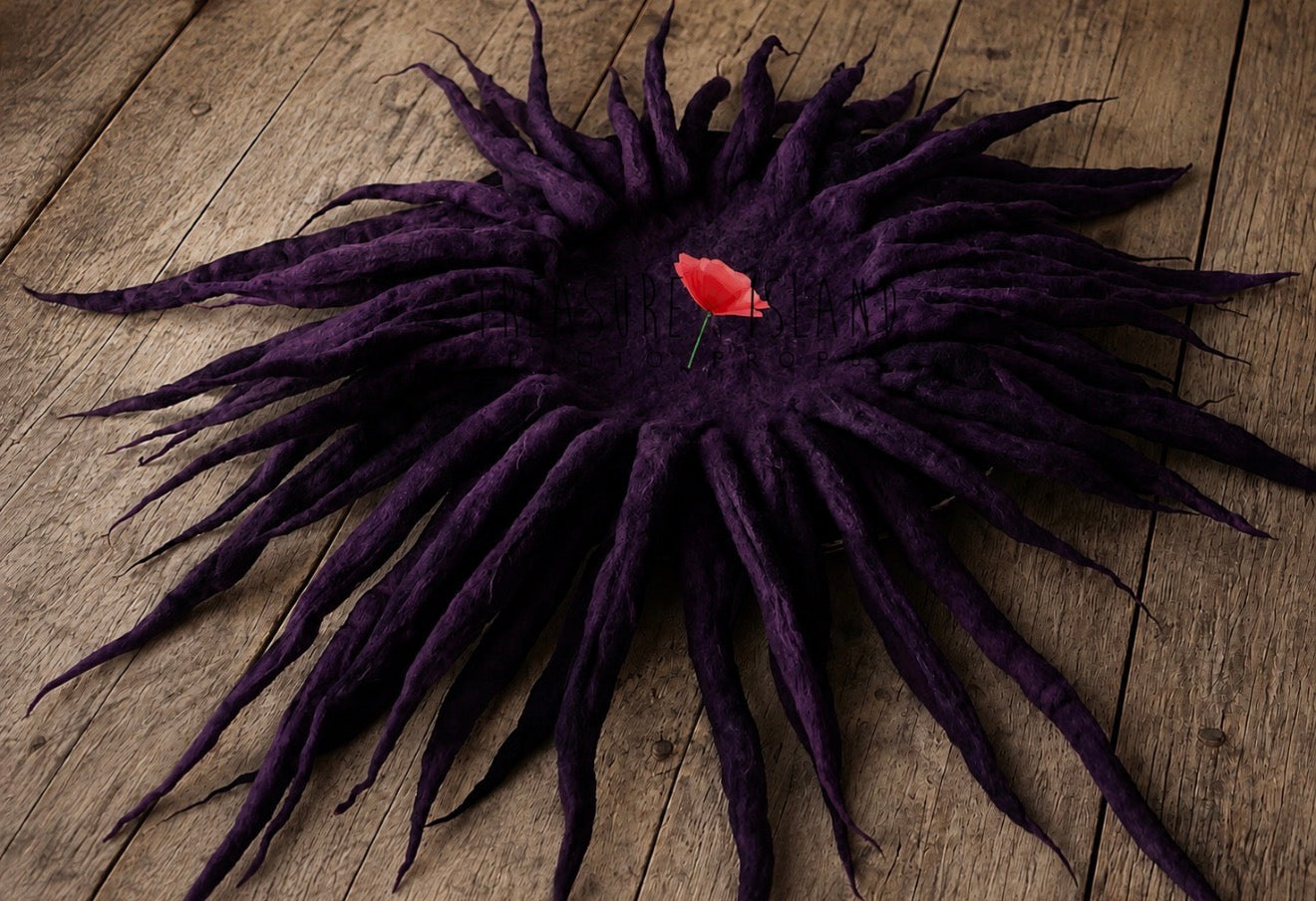 A dark purple felt sunburst hat with long radiating dreadlock-like strands, photographed flat on a rustic wooden surface with a small red flower pin in the center.