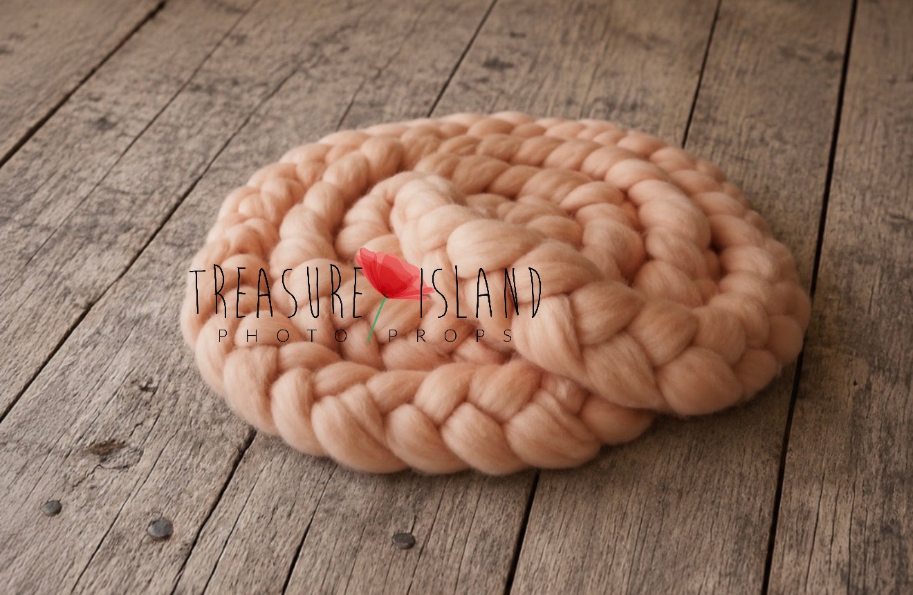 A chunky braided rope pet bed in a soft peachy pink color, coiled on a rustic wooden plank floor. A small red tulip is tucked into the center of the braid.