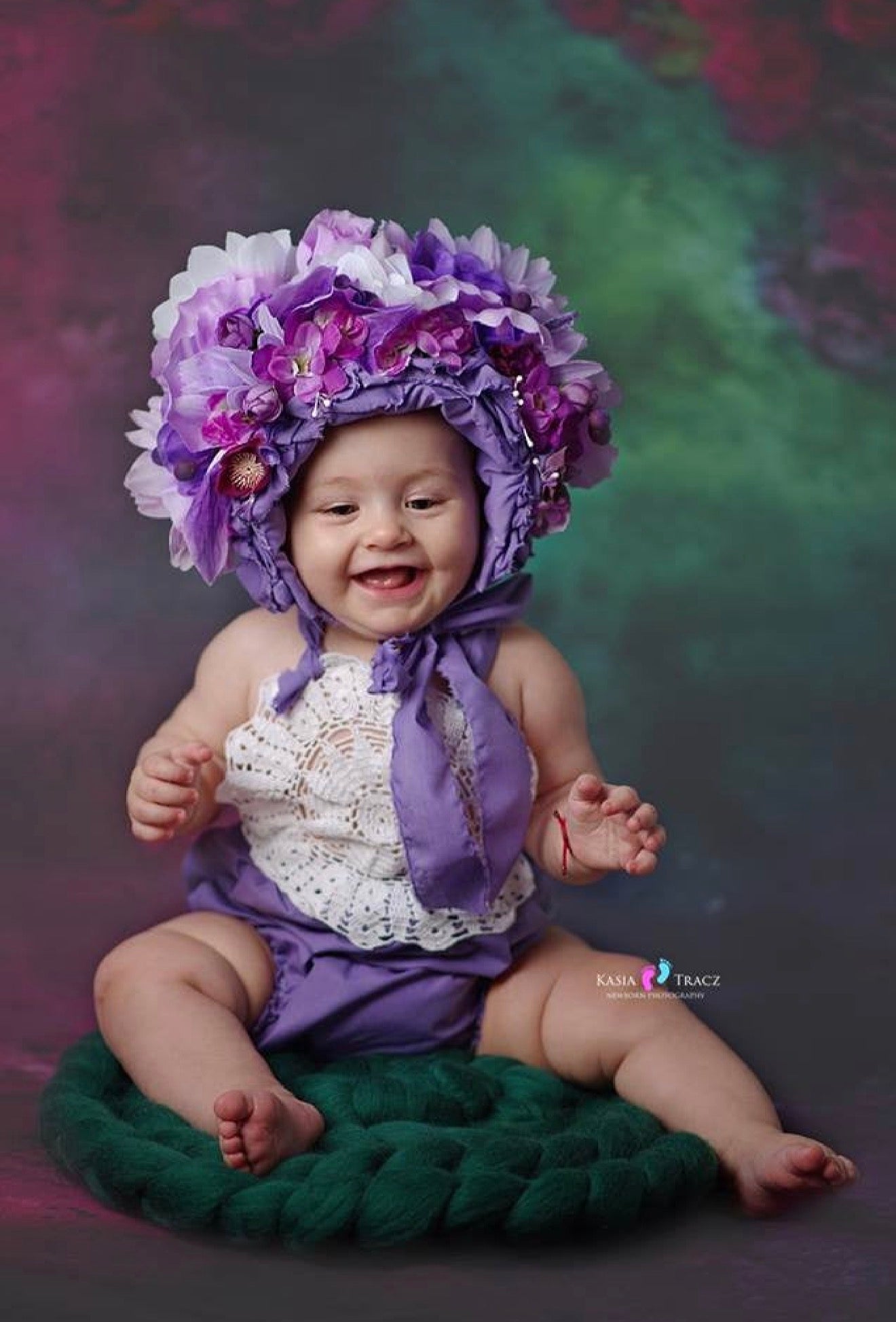 A smiling baby wearing a purple floral bonnet with a purple ribbon and a white lace top, sitting on a green knit cushion against a dark purple and green studio backdrop.