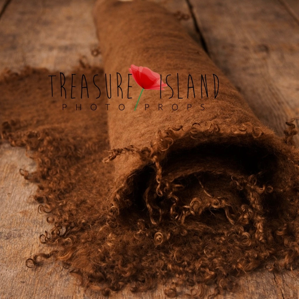 Hand-felted chocolate brown newborn wrap with natural sheep curls, rolled and styled on a light rustic wooden floor – newborn photography prop