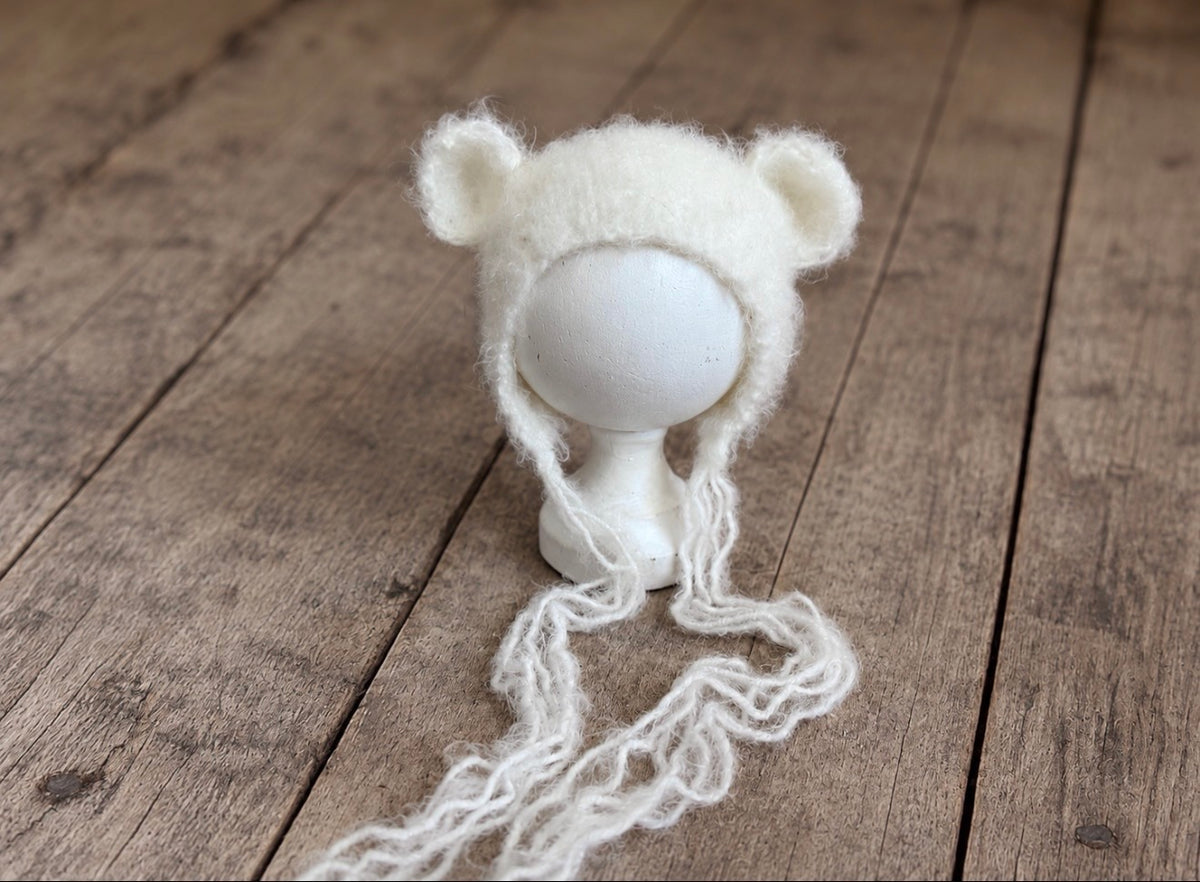 Front view of a fluffy knitted newborn bonnet with rounded teddy bear ears and long soft yarn ties, displayed on a white head form.