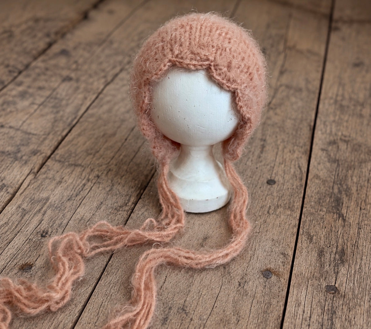 Fluffy dusty pink knitted newborn bonnet with decorative front edging and long ties, displayed on a white mannequin head.