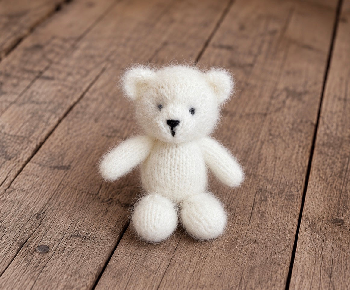 Small handmade knitted teddy bear with rounded ears, black embroidered facial details and a soft fluffy texture, displayed on vintage wooden boards.
