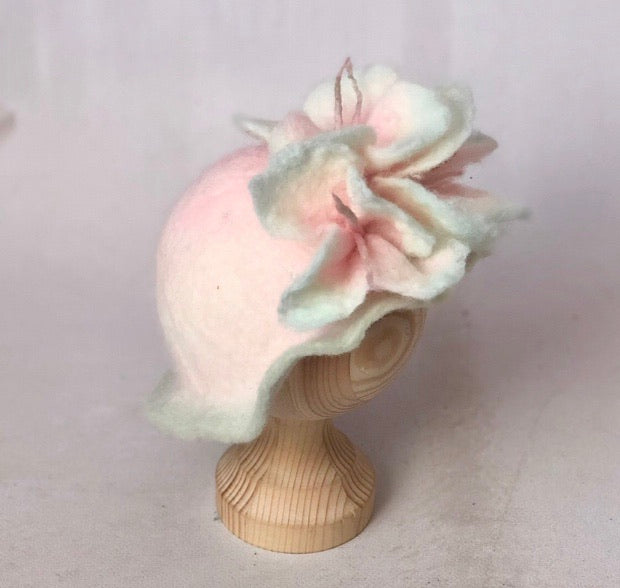 Original studio photograph of a pink hand-felted newborn flower hat with layered pale green petals on a wooden display stand